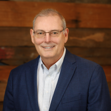 man in light shirt under a dark blue blazer smiling against a wooden wall