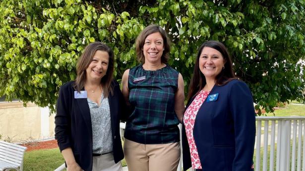Three women stand outside by staircase and green tree smiling.